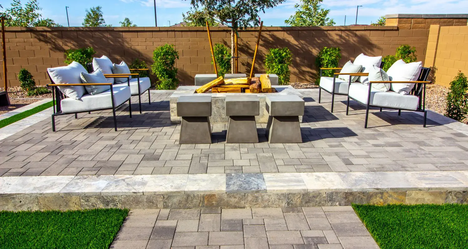 Outdoor patio with gray pavers, seating arranged around a fire pit, and stools. Surrounded by a brick wall and small green shrubs.